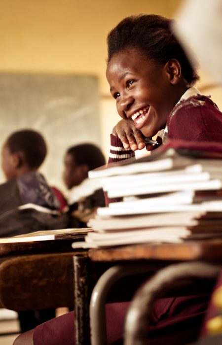 A Happy young South African girl (from the Xhosa tribe) works on her studies and jokes with her friends at at an old worn desk in a class room in the Transkei region of rural South Africa.